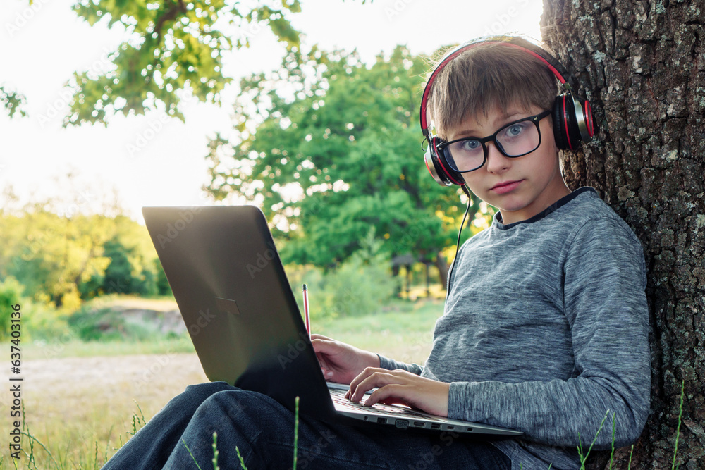 Boy with inquisitive blue eyes in glasses studying while sitting by tree with laptop on lap, listening to task through headphones. Student, surprised by study, taking notes in workbook with pencil. Boy with inquisitive blue eyes in glasses studying while sitting by tree with laptop on lap, listening to task through headphones. Student, surprised by study, taking notes in workbook with pencil.