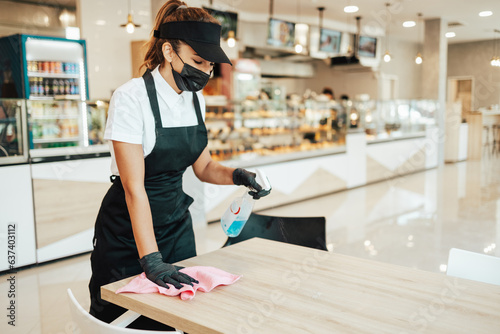 Beautiful woman working bakery or fast food restaurant. She is cleaning and disinfecting tables against Coronavirus pandemic disease. She is wearing protective face masks, gloves and face shield.