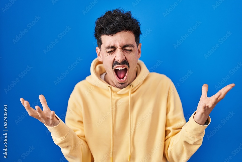 Hispanic man standing over blue background celebrating mad and crazy for success with arms raised and closed eyes screaming excited. winner concept Hispanic man standing over blue background celebrating mad and crazy for success with arms raised and closed eyes screaming excited. winner concept