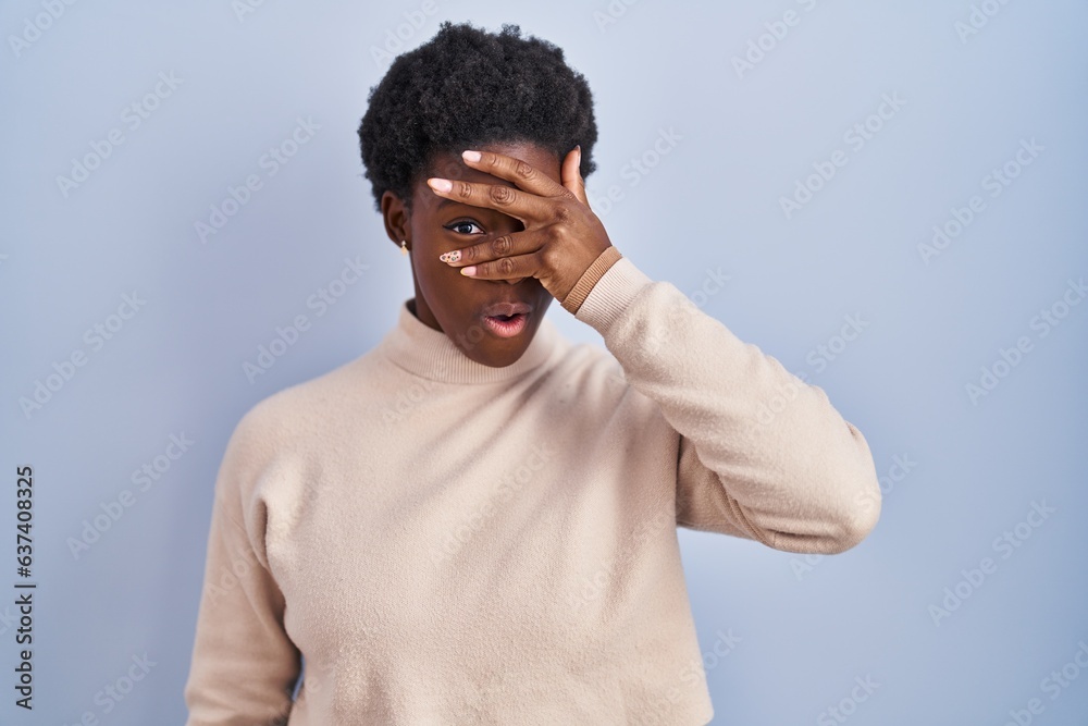African american woman standing over blue background peeking in shock covering face and eyes with hand, looking through fingers with embarrassed expression. African american woman standing over blue background peeking in shock covering face and eyes with hand, looking through fingers with embarrassed expression.