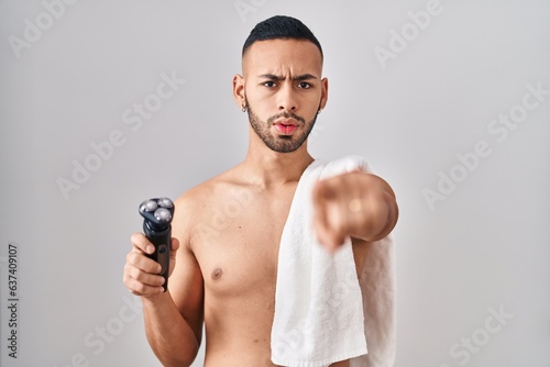 Young hispanic man holding electric razor pointing with finger to the camera and to you, confident gesture looking serious