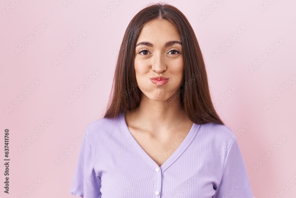 Fototapeta premium Young hispanic woman with long hair standing over pink background puffing cheeks with funny face. mouth inflated with air, crazy expression.
