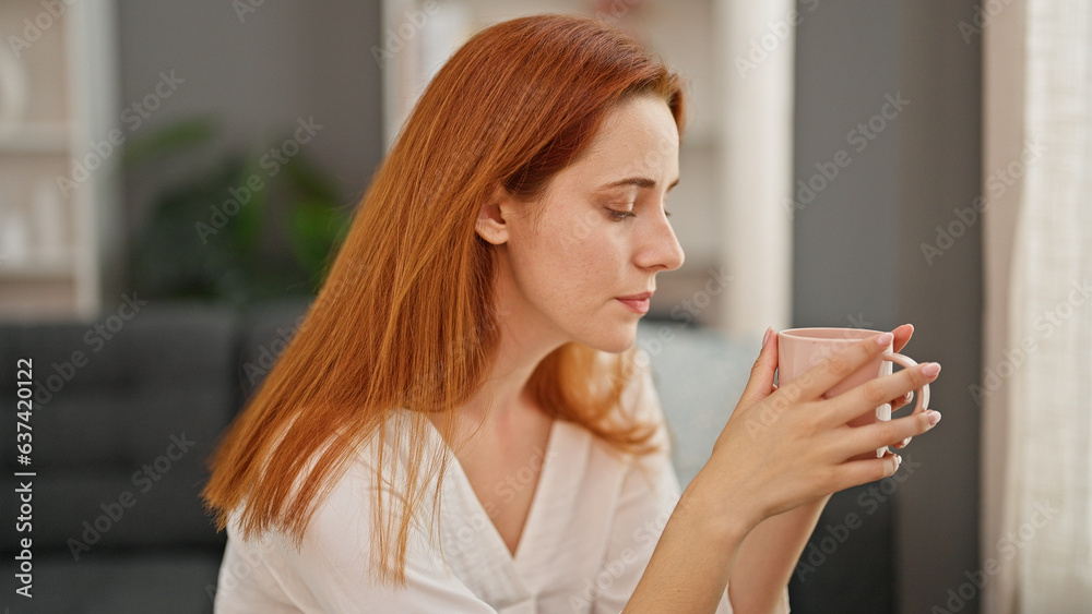 Young redhead woman sitting on sofa with serious expression drinking coffee at home Young redhead woman sitting on sofa with serious expression drinking coffee at home