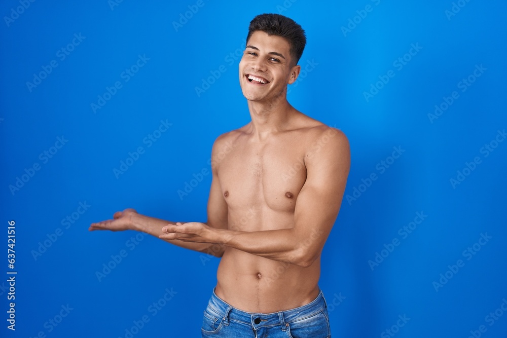 Young hispanic man standing shirtless over blue background inviting to enter smiling natural with open hand