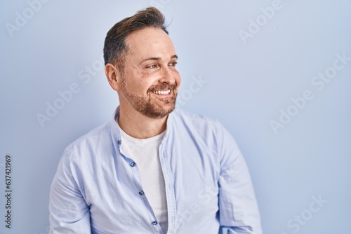 Middle age caucasian man standing over blue background looking away to side with smile on face, natural expression. laughing confident.