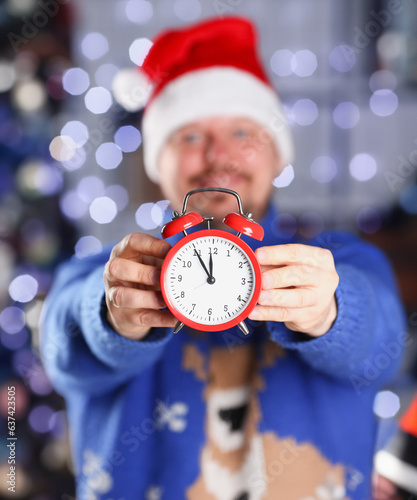 Bearded smiling man wearing warm blue sweater with traditional deer hold arm red alarm clock within inch coming 2024year with glowing garland background concept closeup photo