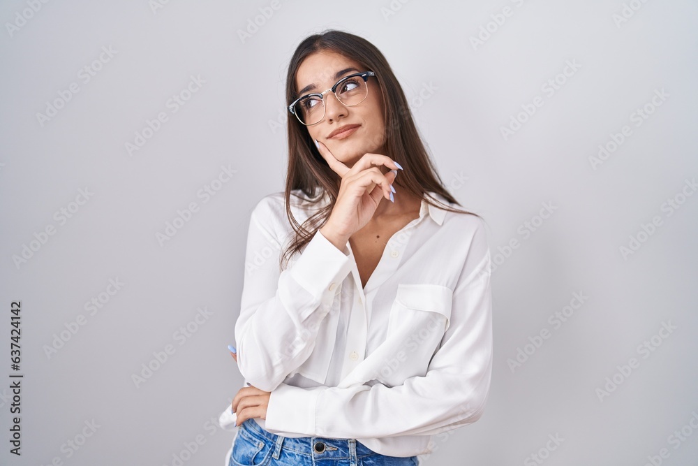 Young brunette woman wearing glasses with hand on chin thinking about question, pensive expression. smiling with thoughtful face. doubt concept. Young brunette woman wearing glasses with hand on chin thinking about question, pensive expression. smiling with thoughtful face. doubt concept.