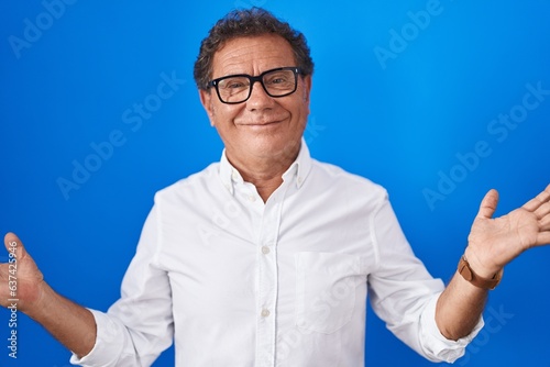 Middle age hispanic man standing over blue background clueless and confused expression with arms and hands raised. doubt concept.