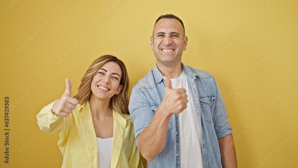Man and woman couple standing together doing thumb up gesture over isolated yellow background Man and woman couple standing together doing thumb up gesture over isolated yellow background