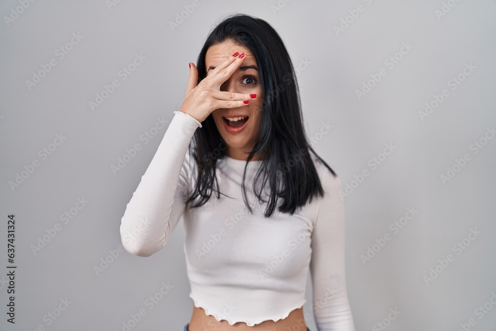 Hispanic woman standing over isolated background peeking in shock covering face and eyes with hand, looking through fingers with embarrassed expression. Hispanic woman standing over isolated background peeking in shock covering face and eyes with hand, looking through fingers with embarrassed expression.