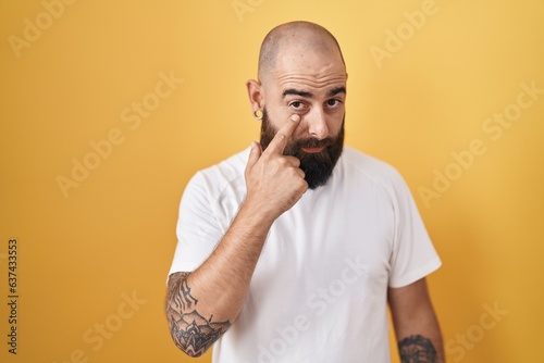 Young hispanic man with beard and tattoos standing over yellow background pointing to the eye watching you gesture, suspicious expression