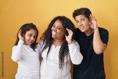 Family of mother, daughter and son standing over yellow background smiling with hand over ear listening an hearing to rumor or gossip. deafness concept.