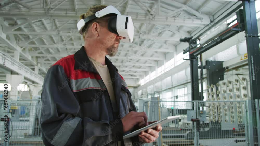 Medium shot of male engineer wearing VR headset standing in industrial factory and working on digital tablet