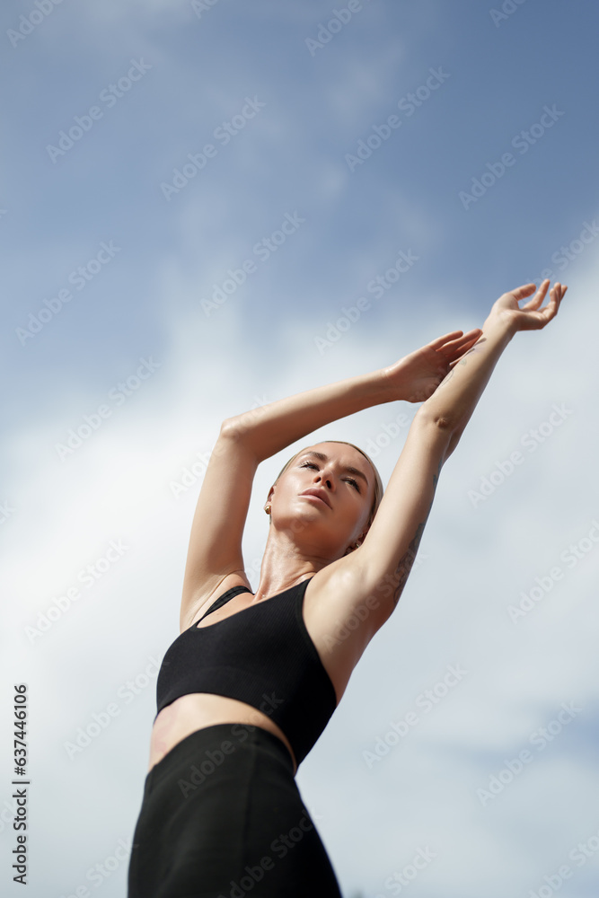 A blonde fitness model in black sportswear stretching her arms up towards the sky. She is standing against a blue sky with white clouds A blonde fitness model in black sportswear stretching her arms up towards the sky. She is standing against a blue sky with white clouds
