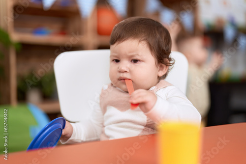 Adorable toddler sitting on table sucking plastic spoon holding dish at kindergarten