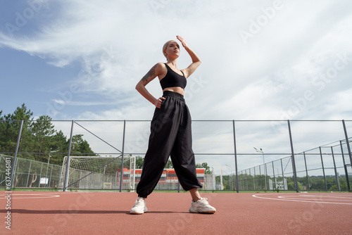A photo of a blonde fitness model with a short haircut posing confidently on a red playground. She is wearing a black crop top and black baggy pants with white sneakers © arthurhidden