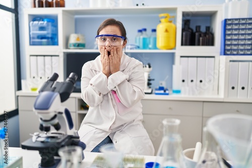 Hispanic girl with down syndrome working at scientist laboratory afraid and shocked, surprise and amazed expression with hands on face