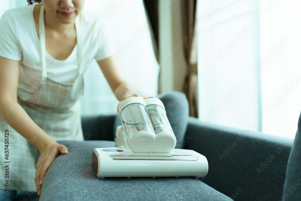 Fototapeta premium Asian housewife using a vacuum machine to clean a sofa in living room close up. Asian housekeeper vacuuming in living room. Chores and daily housework activities concept.