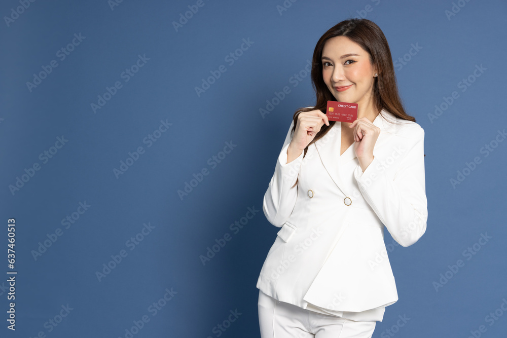 Young beautiful Asian business woman in white suit smiling holding credit card isolated on blue background Young beautiful Asian business woman in white suit smiling holding credit card isolated on blue background