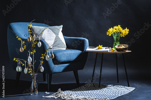 interior, holidays and home decor concept - modern blue chair with pillow, easter eggs hanging on forsythia branches in vase on floor and daffodil flowers on table in dark room