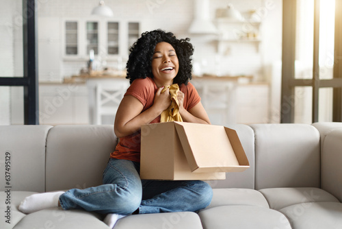 фотографія Excited Young African American Female Unpaking Box With New Clothes At Home