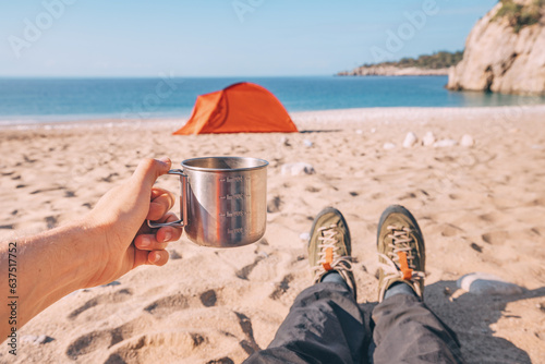 Metal cup with hot tea against background of orange tent on a sandy beach during coastal hike
