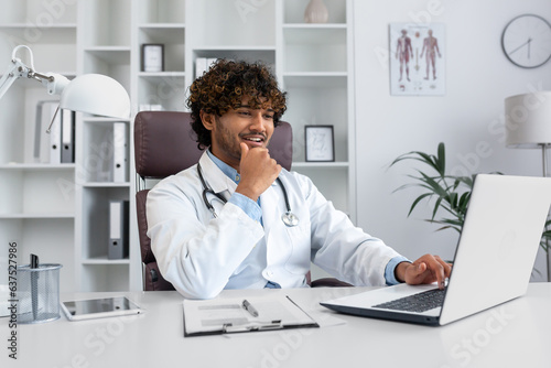 An Indian smiling young doctor in a white coat sits in the office at a table and works on a laptop, talks on the phone with a patient, consults.