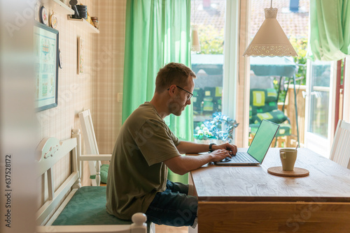 A man in glasses sitting at the computer indoors in a private house filled with sunlight. Freelancer, workation, work at home. 