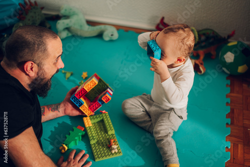 A fun dad is playing a game with his silly toddler while holding a plastic brick toy and babysitting a child. photo