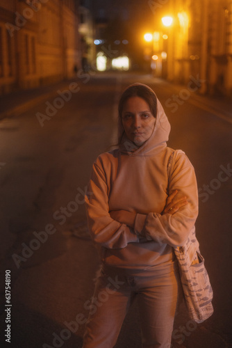 young thoughtful woman stands on road in beige hoodie. walk in evening in light of lanterns, portrait of female alone. freedom loneliness of modern generation. Travel and rest alone, mental health