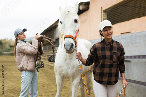 Women ranchers preparing white horse for ride. Asian woman leading horse, European woman carrying saddle.