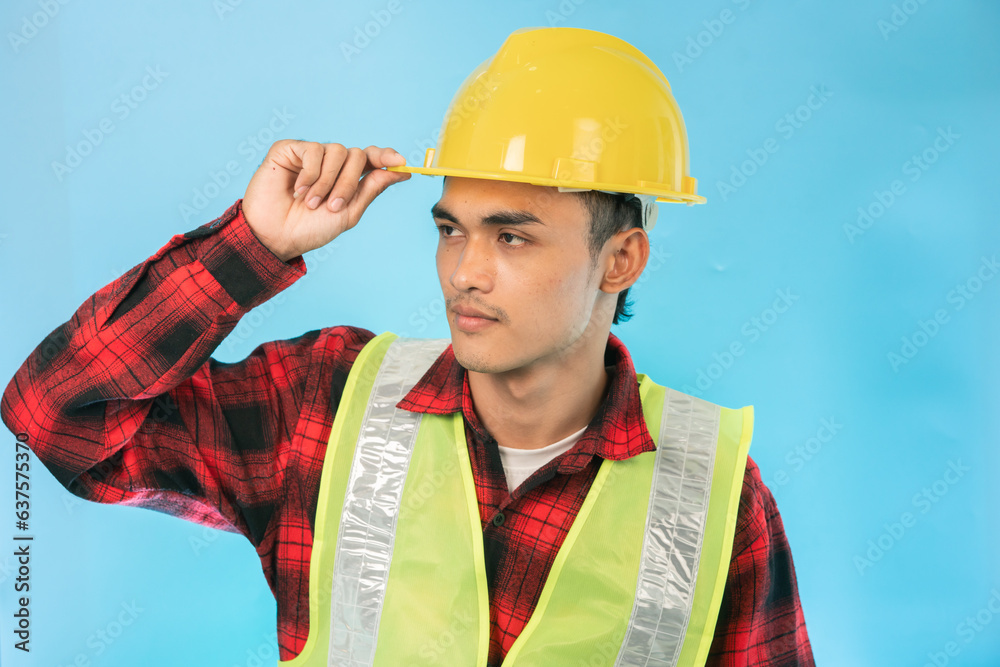 A young engineer wearing a yellow protective helmet, holding the helmet, looking determined. Isolated on a blue background A young engineer wearing a yellow protective helmet, holding the helmet, looking determined. Isolated on a blue background