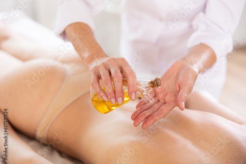 Professional massage therapist applying natural oil to her hands to make back massage to young female client more comfortable, nourishing skin, promoting relaxation and providing aromatherapy benefits
