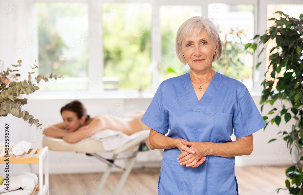 Obraz premium Successful confident mature female massage therapist standing in procedure room of modern wellness center, looking at camera with smile..