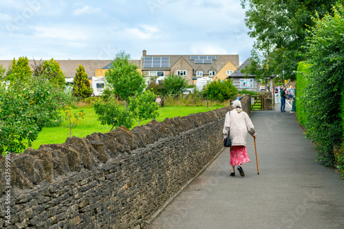 A senior woman with a cane walks alone down an alley next to a stone wall in the picturesque Cotswolds village of Bourton-on-the-Water, England, United Kingdom. photo