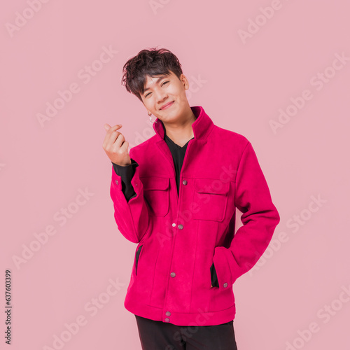 Portrait of a happy Asian handsome man in fashionable pink jacket clothing standing smiling and using fingers to make a mini heart sign isolated on pink background.