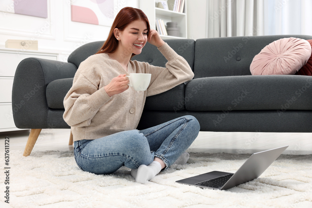 Happy woman with cup of drink and laptop on rug in living room