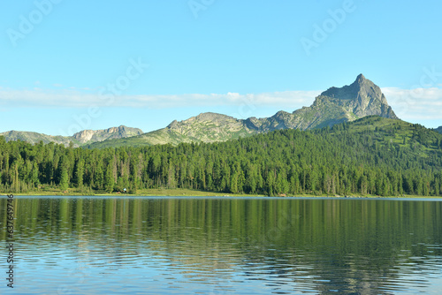 Reflection of a high rock on the unsteady surface of a large lake in a coniferous forest on the shores on a sunny summer day.