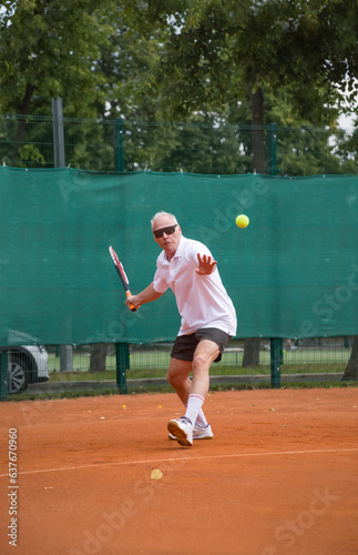 An elderly man playing tennis is hitting a forehand. The moving at camera. Open ground.