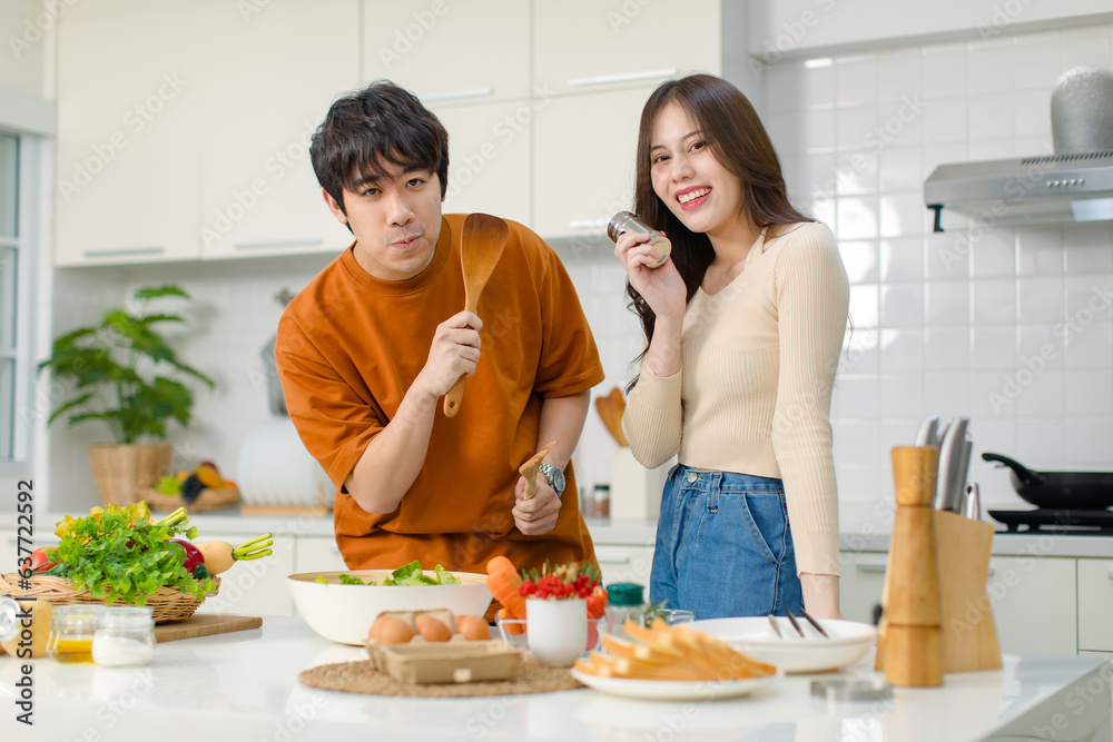 Asian young lovely lover couple husband and wife in casual outfit standing smiling holding red tomato while mixing vegetable salad with wooden fork and spoon in bowl in full decorated modern kitchen