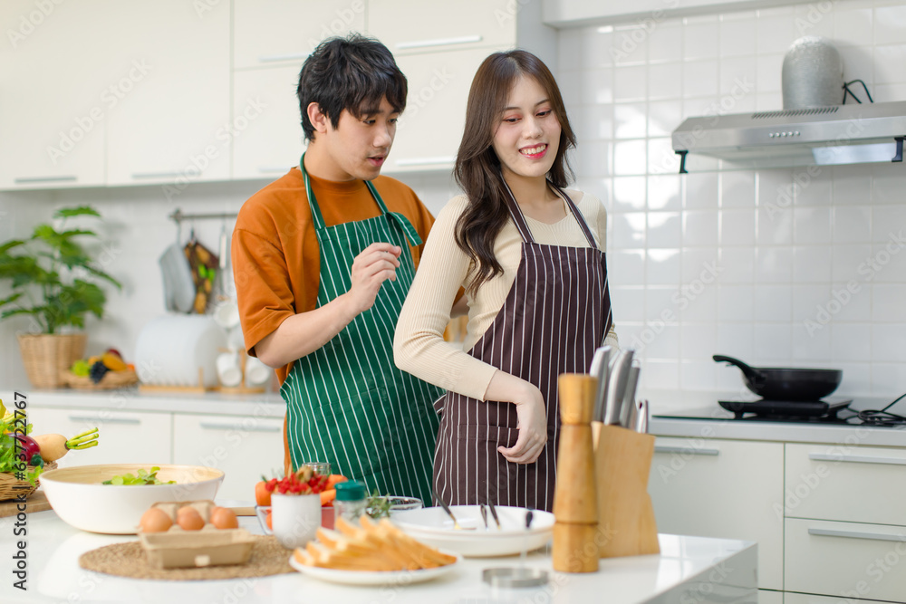 Asian young lovely lover couple husband and wife in casual outfit standing smiling male helping wearing clean striped apron to female in full decorated modern kitchen with cooking equipment at home