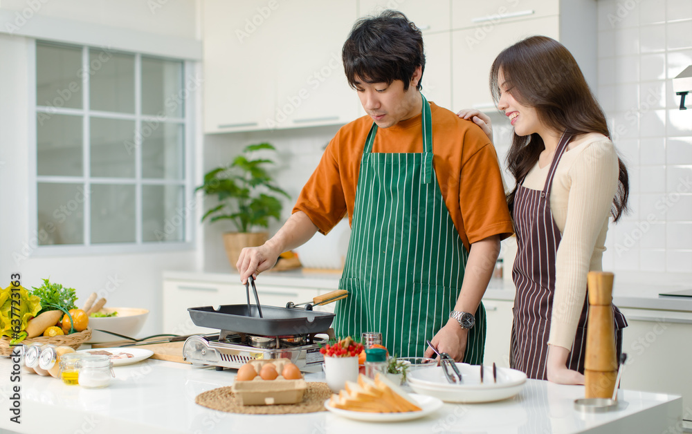 Asian young lover couple husband and wife in casual outfit with apron standing smiling while man cooking frying food with pan woman helping in full decorated modern kitchen with ingredients equipment