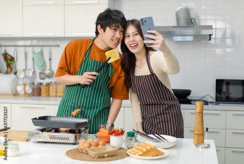 Asian young lover couple husband and wife in casual outfit with apron standing smiling taking selfie photo with smartphone together while man holding bread in full decorated modern kitchen at home
