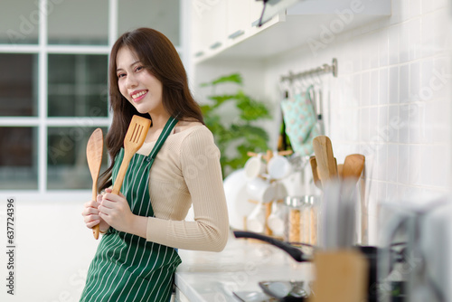 Asian young beautiful housewife in casual outfit with apron standing smiling holding wooden spoon and fork utensil preparing to frying in cooking pan in full decorated modern kitchen with equipment