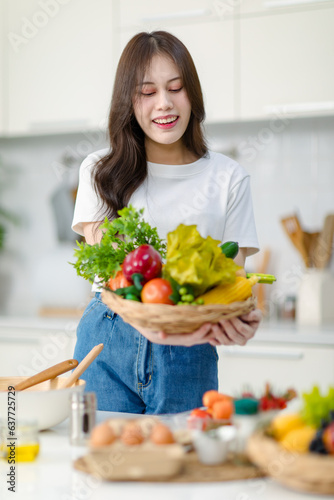 Asian young beautiful housewife in casual outfit standing smiling showing red sweet pepper and mixed fresh organic vegetables basket in full decorated modern kitchen with ingredients and equipment