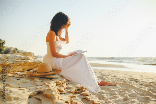 A girl in a long white dress straightens her hair while reading a book while sitting on a stone near the sea, lake or ocean.