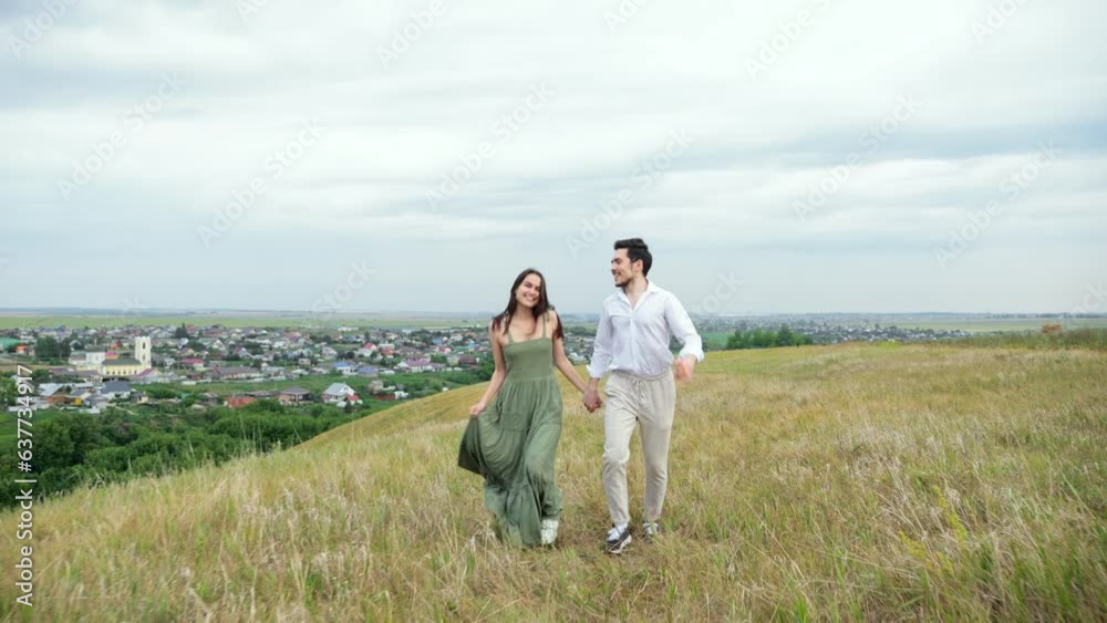 Positive couple of man and woman run across hilly field against huge town. Concept of view of territory with buildings and love slow motion Positive couple of man and woman run across hilly field against huge town. Concept of view of territory with buildings and love slow motion