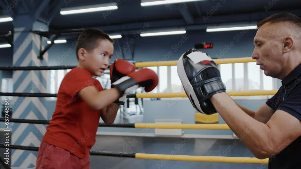 Man coach trains little boy in boxing gloves to box using boxing paws, side view, gym. Father trains with his son in martial art at gym Man coach trains little boy in boxing gloves to box using boxing paws, side view, gym. Father trains with his son in martial art at gym