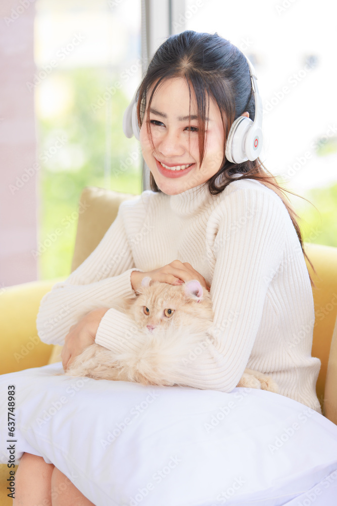 Asian young beautiful female teenager girl owner in turtleneck sweater wearing headphones listening to music sitting smiling on cozy sofa holding cute little orange long hair domestic cat on pillow Asian young beautiful female teenager girl owner in turtleneck sweater wearing headphones listening to music sitting smiling on cozy sofa holding cute little orange long hair domestic cat on pillow