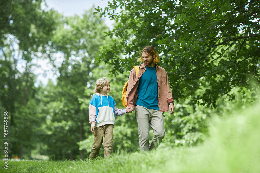 Low angle portrait of happy modern father and son holding hands while walking on green grass in park together, copy space Low angle portrait of happy modern father and son holding hands while walking on green grass in park together, copy space